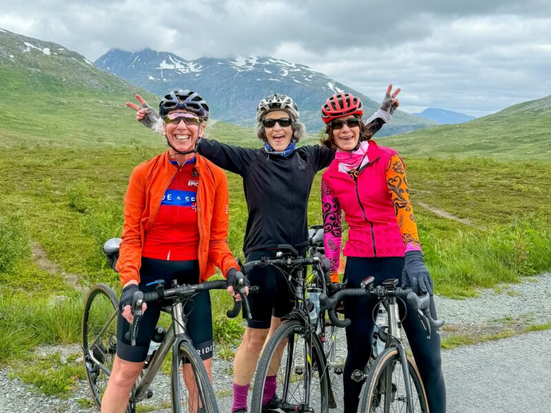 Three smiling cyclists in colourful jerseys pose with their bikes with snow-capped peaks behind them.