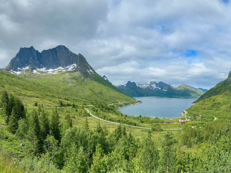 A panoramic view of a fjord surrounded by steep green mountains, with scattered houses along the water’s edge.