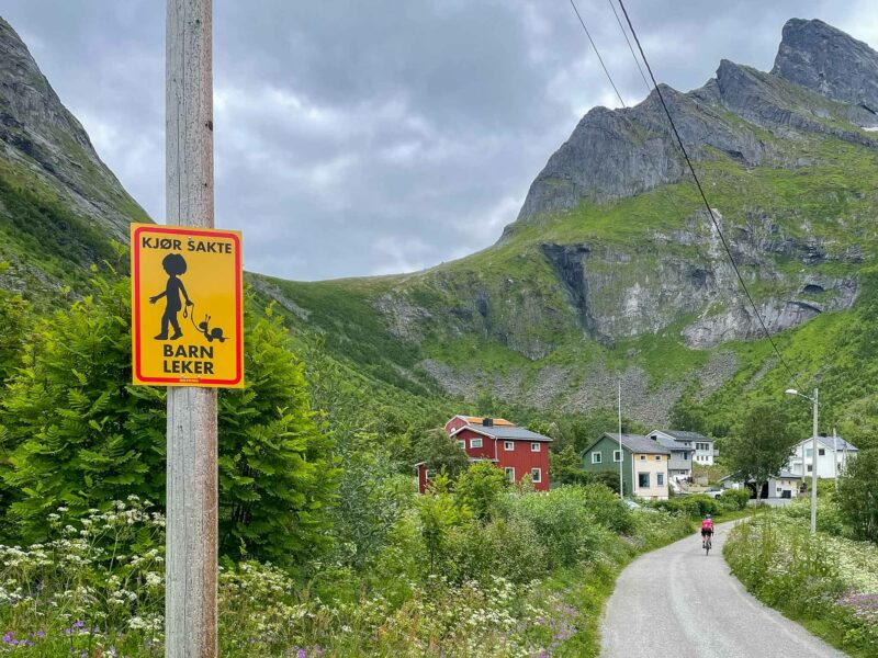 Yellow road sign with a child and toy warning drivers to slow down, with green mountains and houses in the background.