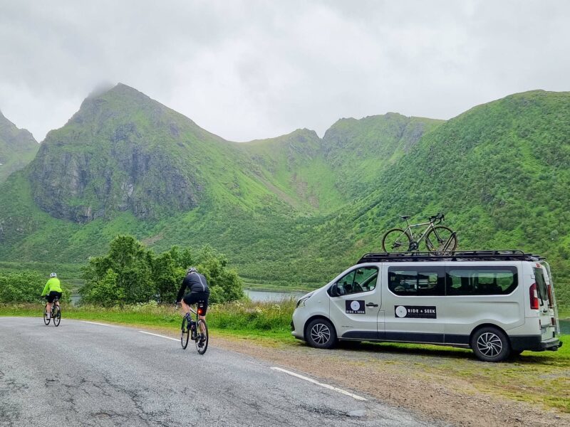 Two cyclists riding past a support van with bikes on the roof, surrounded by misty green mountains.