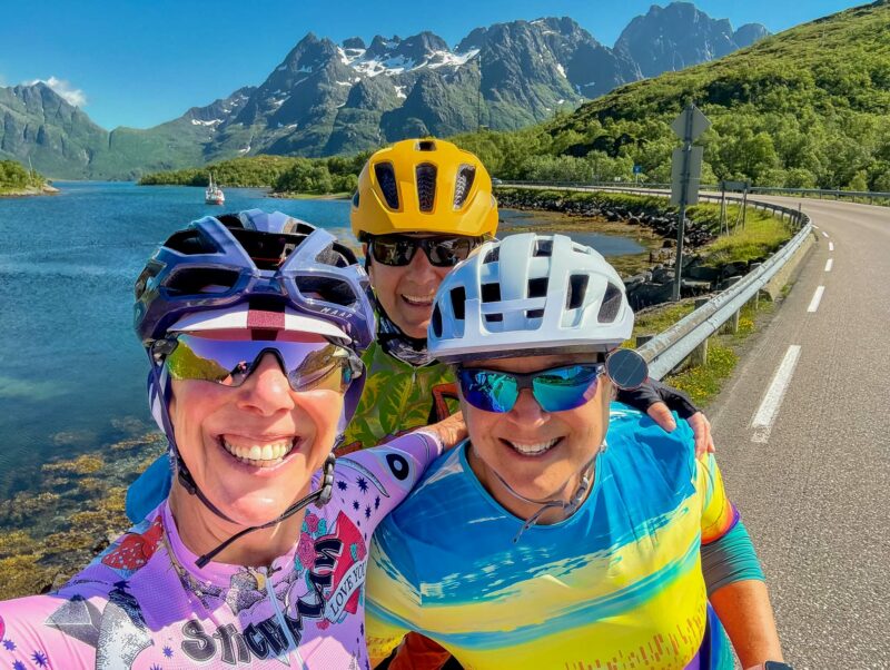 Three smiling cyclists in colourful jerseys taking a selfie on a road by a fjord, with sharp mountain peaks behind them.