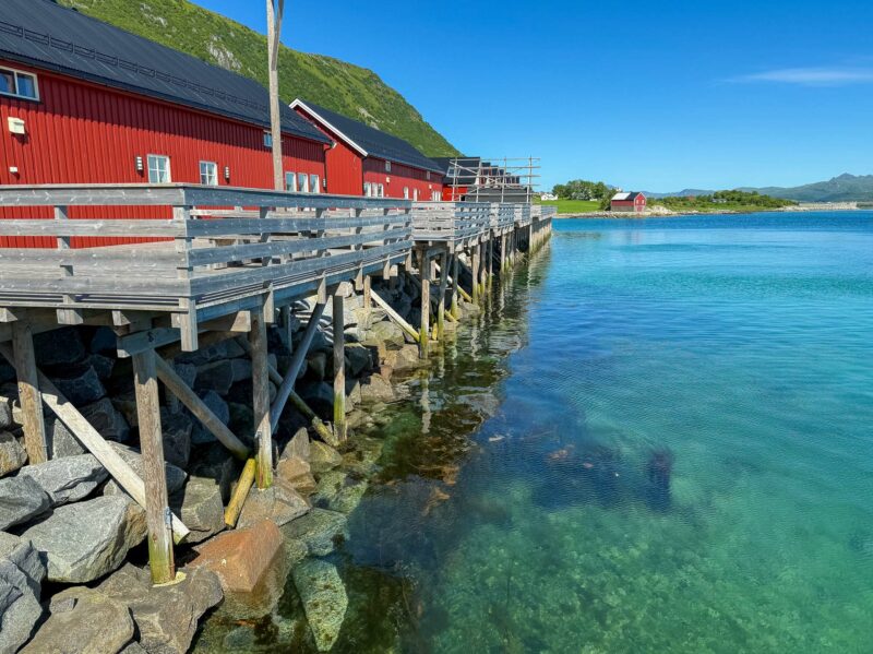 Traditional red wooden cabins on stilts above clear turquoise water along a rocky shoreline.