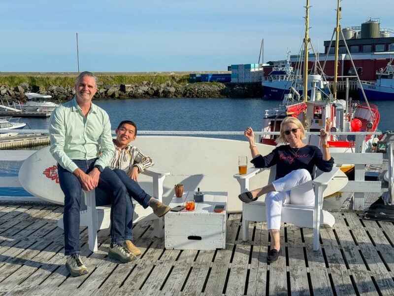 Three people sitting relaxing with drinks seated on white chairs at a harbor dock with fishing boats in the background.