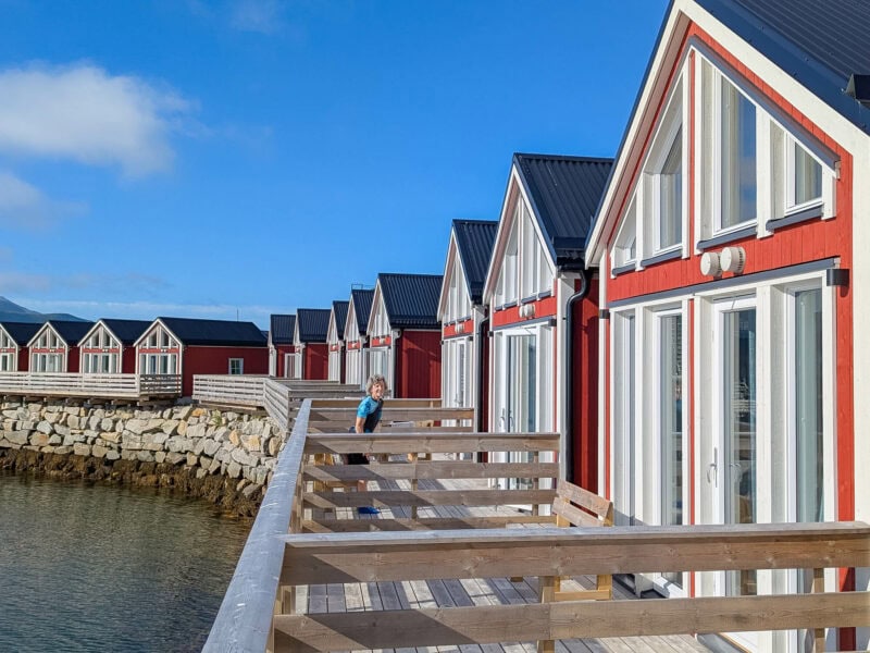 A line of red and white waterfront cabins with large windows, reflecting in the calm water under a bright blue sky. A cyclist can be seen standing on one of the balconies.