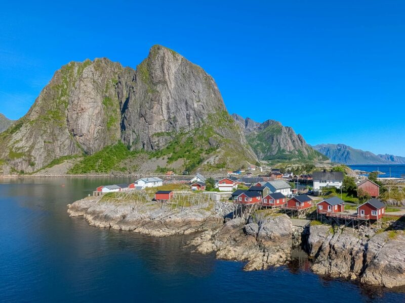 A small coastal village with red cabins perched on rocky shores, backed by towering green cliffs.