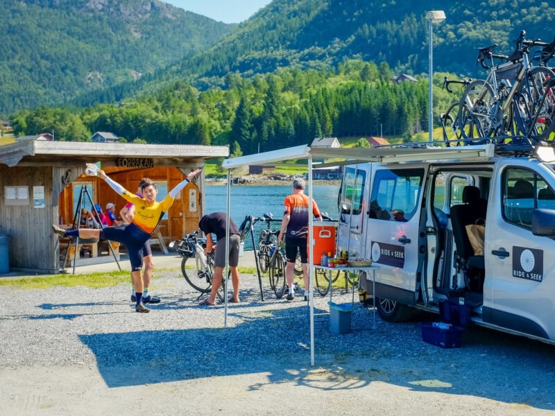 A cheerful cyclist in a yellow jersey posing by a Ride & Seek support van while a guide works on bikes and other cyclists are seated in a nearby shelter near a fjord.