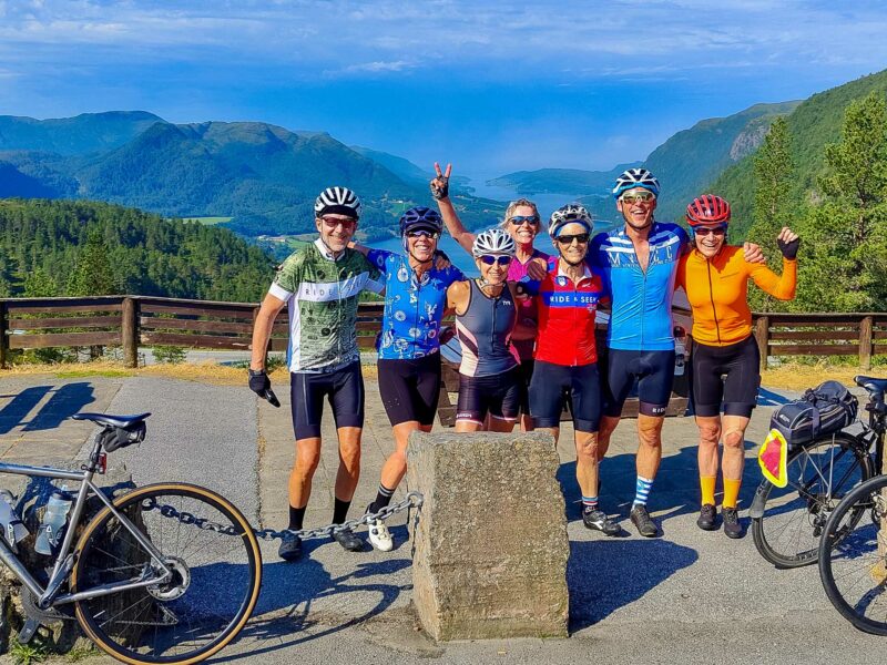 A group of cyclists in colourful jerseys posing together at a scenic overlook with fjords and forested hills in the background.