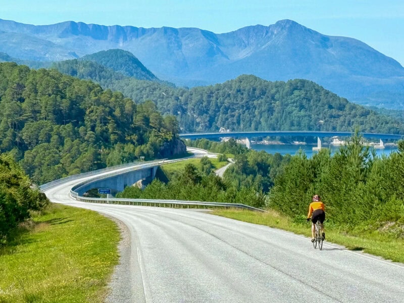 A lone cyclist in an orange jersey riding along a winding fjordside road toward a distant bridge with mountains in the background.