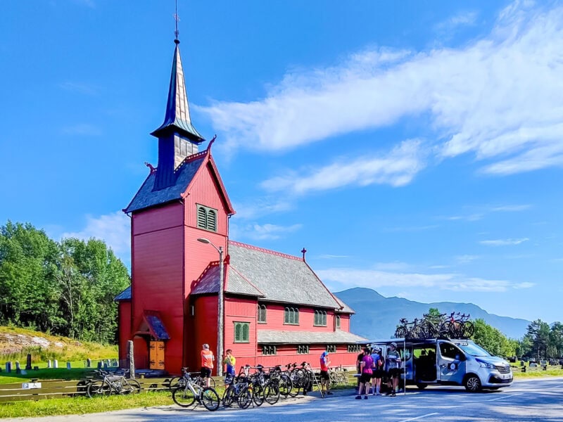 Cyclists and a Ride & Seek support van parked outside a small red wooden church with a tall steeple with cyclists for a snack break.