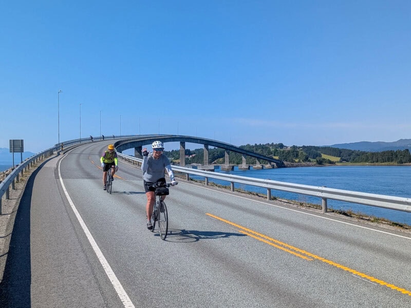 Two cyclists ride across a sweeping bridge over the fjord, smiling and waving under a clear blue sky.