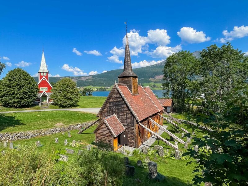 A medieval wooden stave church with braced walls next to a red chapel, set in a green valley by a lake.