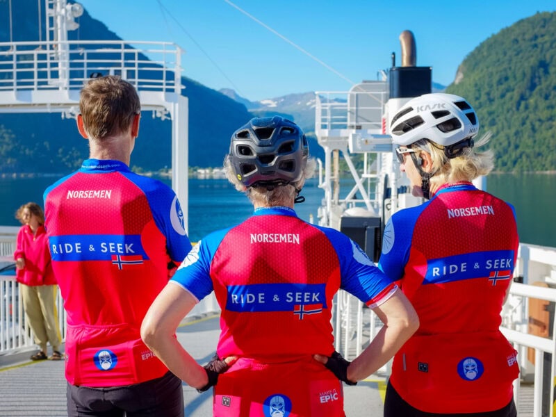 Three riders in matching red "Ride & Seek" jerseys stand with their backs to the camera, gazing over the water and surrounding fjords.