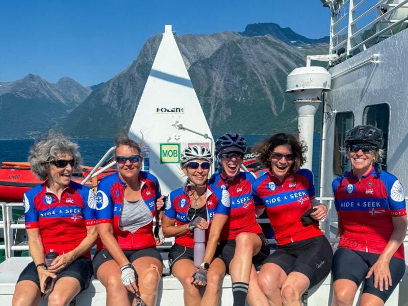 Six cyclists in matching red Ride and Seek jerseys sit together on the deck of a ferry, laughing with fjord cliffs and blue water behind them.