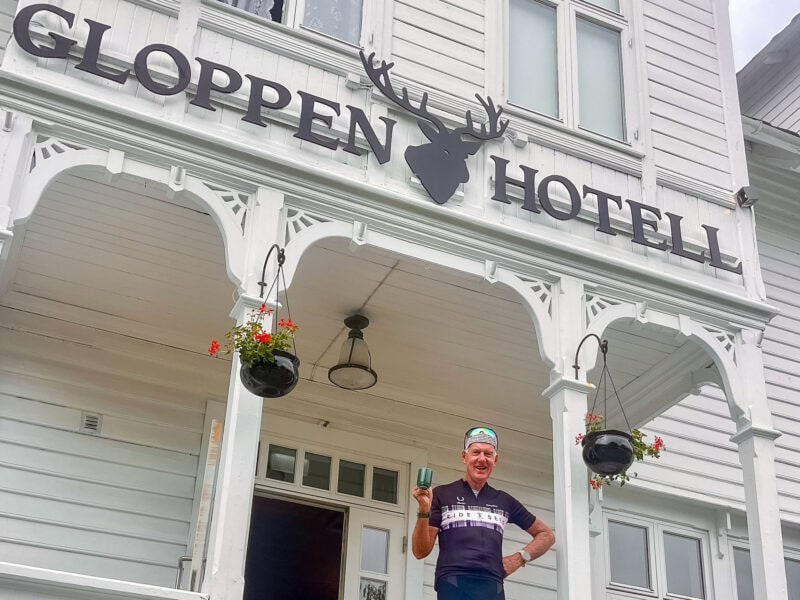 A smiling cyclist stands holding a drink in front of the historic white wooden Gloppen Hotell building with decorative trim.