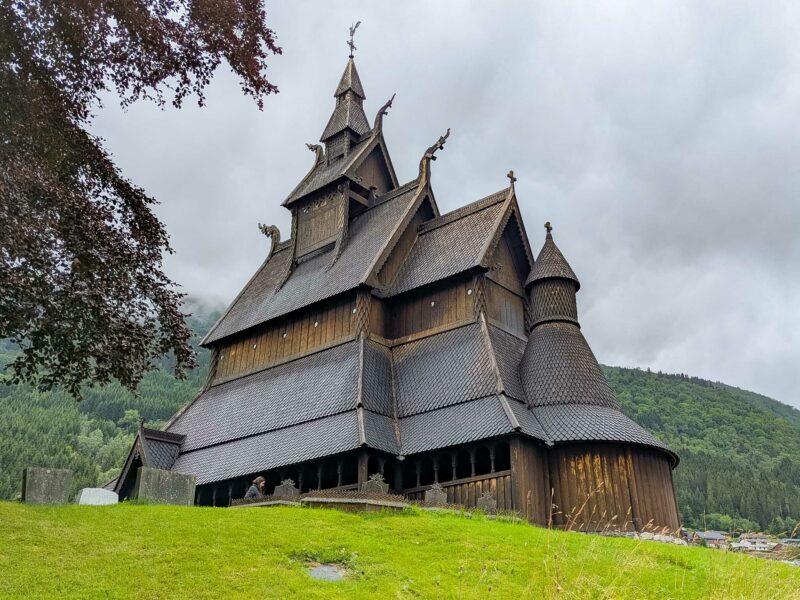A dark wooden Norwegian stave church with steep gables and dragonhead carvings stands on a grassy hill under cloudy skies.