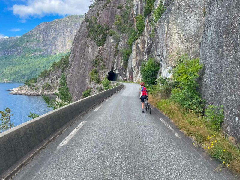 A Cyclist in red poses along a narrow coastal road carved into cliffs, with a tunnel ahead and blue fjord waters beside them.