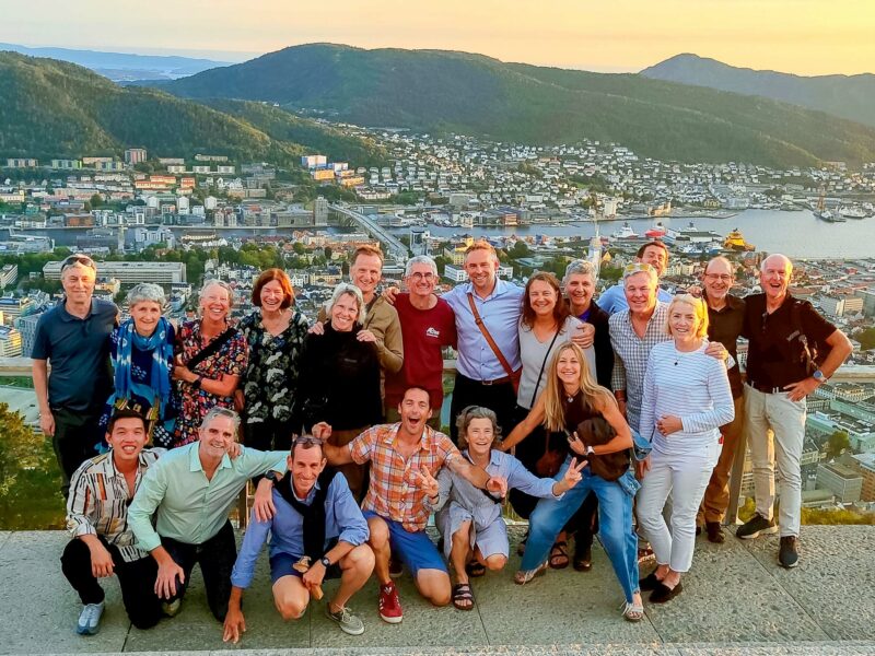 Whole tour group poses on a viewpoint above Bergen at sunset, with the city, harbor, and surrounding mountains below.