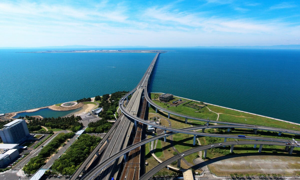 A small island in the background is joined by a long straight bridge. A large expanse of blue water divides the island from the headland.
