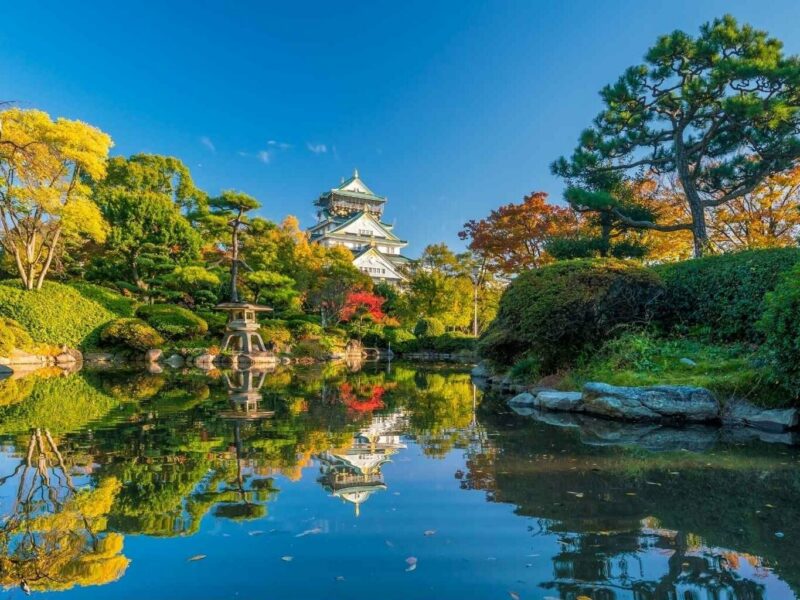 A still lake surrounded by lush gardens. A white, traditional temple sits amongst the gardens in the background.