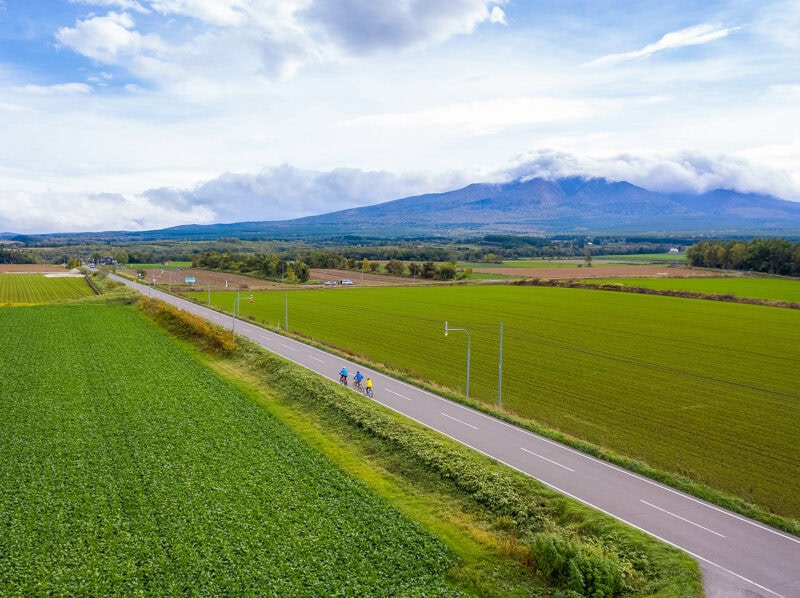 Three cyclists riding a rural road bordered by green farmland, with distant mountains under a cloudy sky.