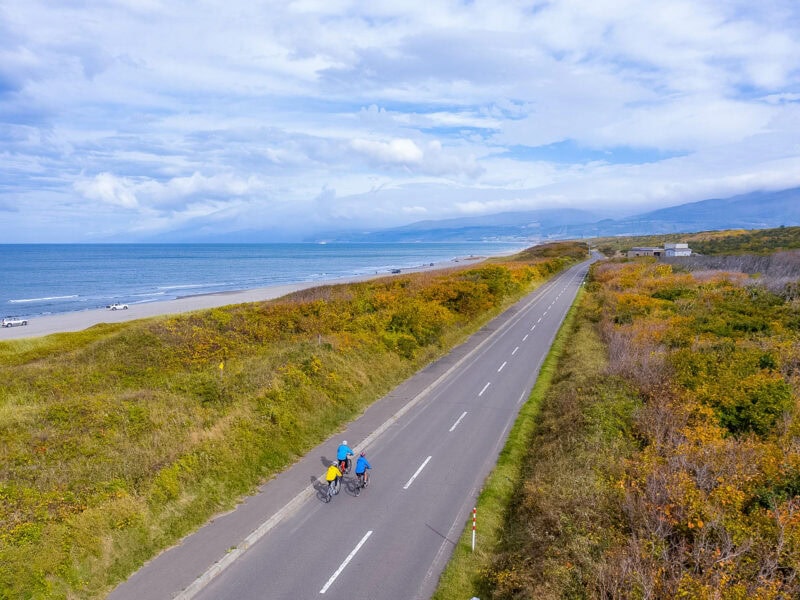Pair of cyclists riding along a coastal highway with the sea on one side and grassy slopes on the other. Mountains stretching away from the coastline in the background.