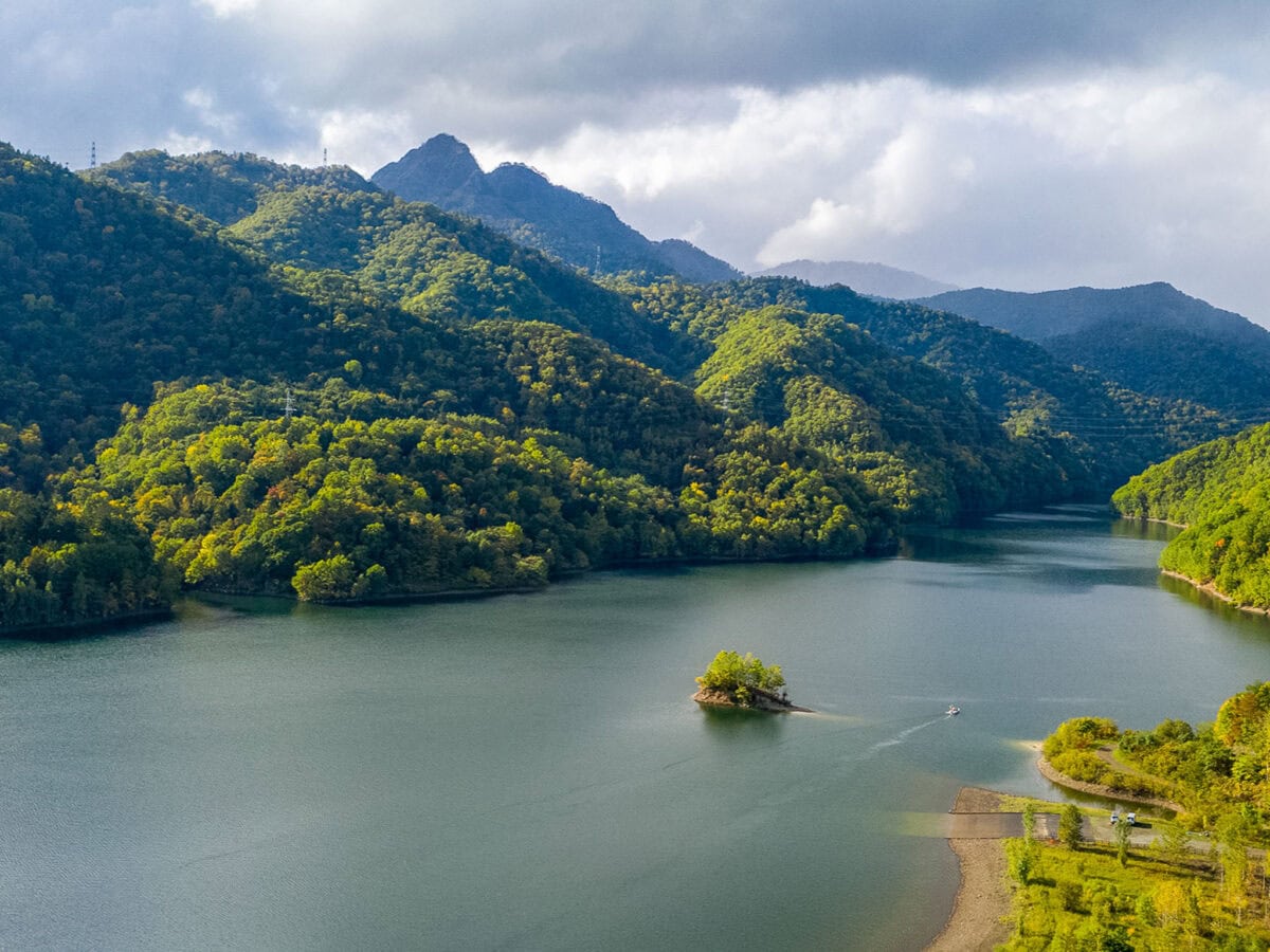 Tranquil lake bordered by forested hills and mountains, with soft sunlight highlighting the green forest and grey clouds drifting across the sky.