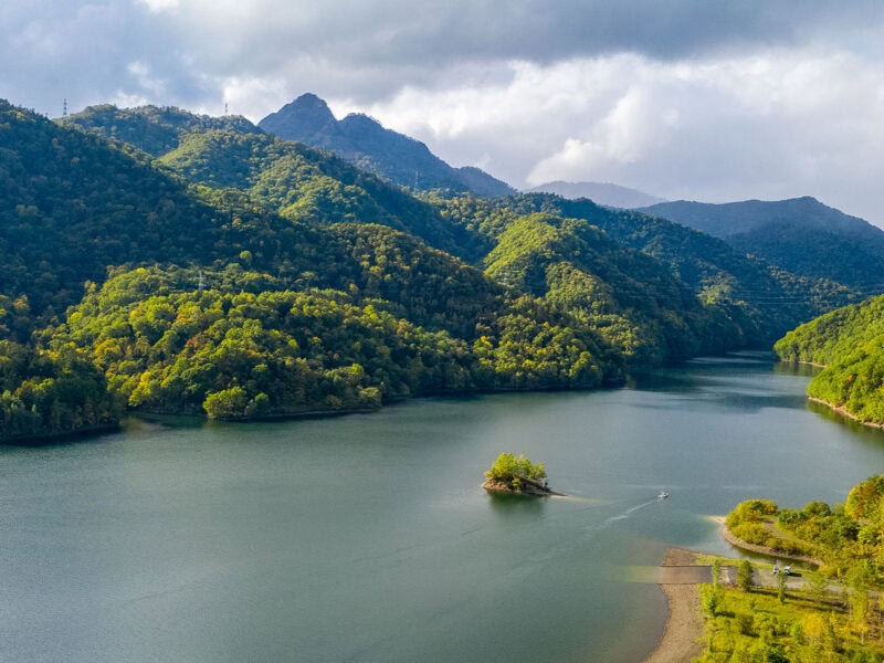 Tranquil lake bordered by forested hills and mountains, with soft sunlight highlighting the green forest and grey clouds drifting across the sky.