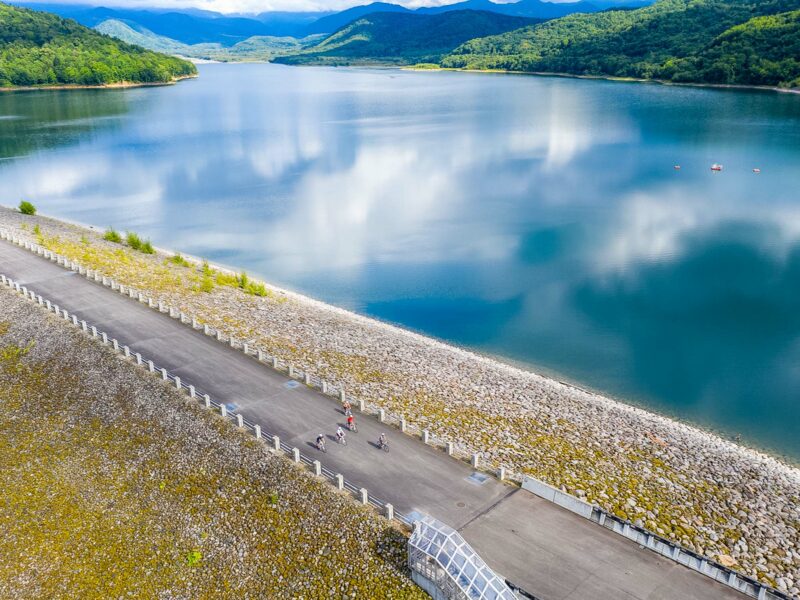 Expansive calm reservoir reflecting clouds and hills, with dam embankments in the foreground.