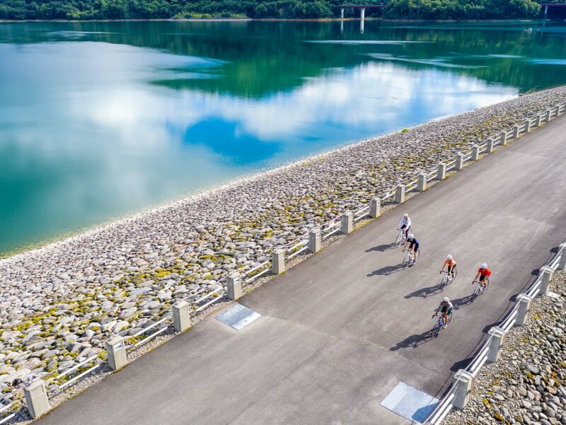 Group of cyclists riding across a wide paved dam road, bordered by stone embankments with turquoise reservoir water beside them.