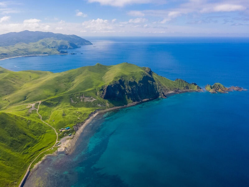 Grassy peninsula extending into a bright blue sea, with steep cliffs and multiple small bays visible along the coastline.