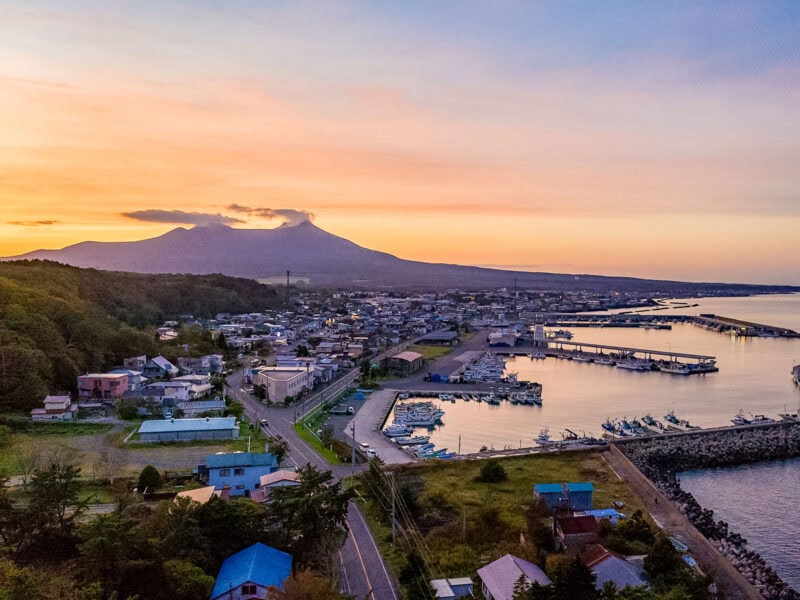 Fishing town with docks and houses along the shoreline, backed by a volcanic peak glowing in soft pink and orange sunset light.
