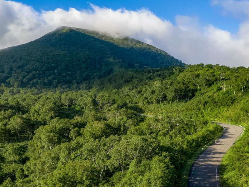 Steep green mountain capped by a low cloud near its summit, with two cyclists riding along a winding road at the forested base.