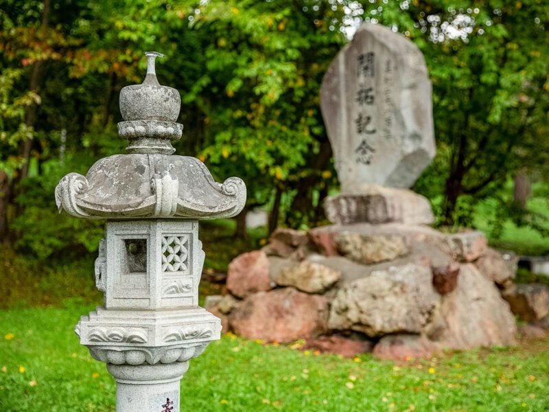 Traditional carved stone lantern standing in front of a large engraved monument, set on a base of boulders in a grassy garden with trees behind.