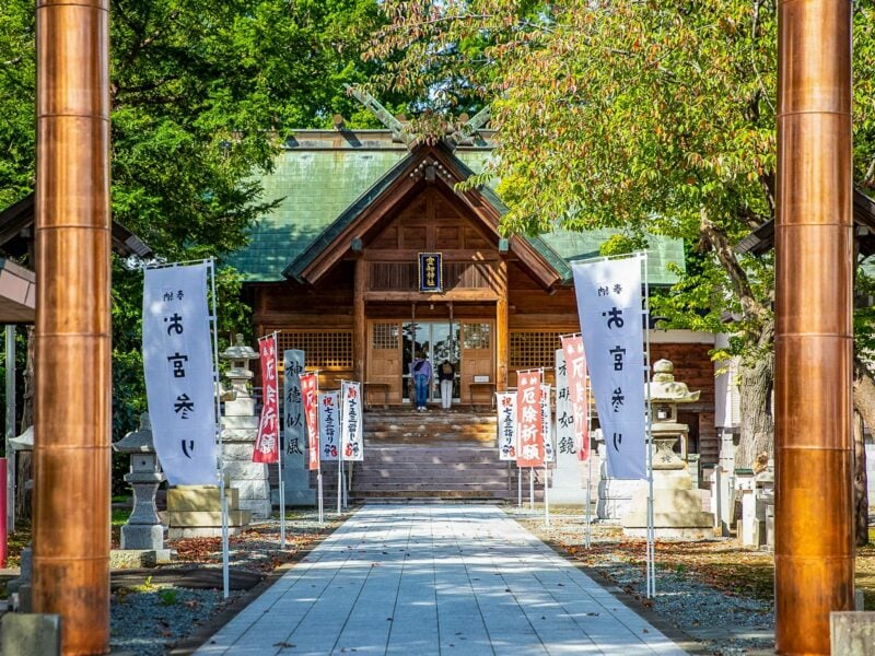 Wooden shrine with green roof and banners, framed by tall trees along a stone pathway lined with flags and traditional stone statues. Two copper pillars frame the scene in the foreground.