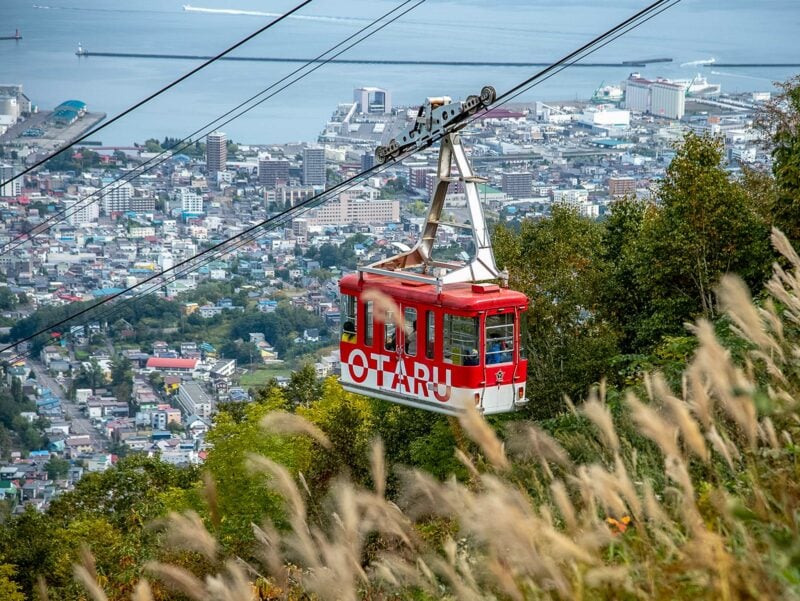 Red gondola marked “Otaru” ascending above grassy hillside, with a cityscape and harbor visible in the background.