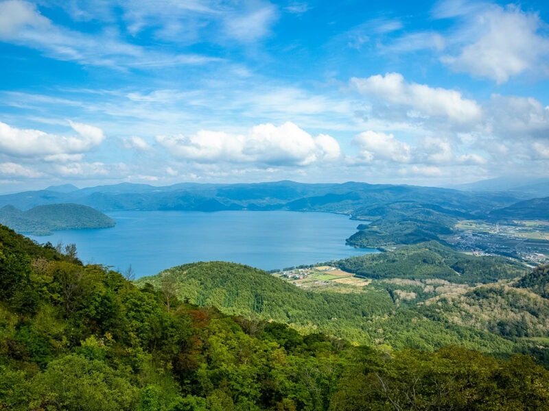 Expansive view over a deep blue lake surrounded by rolling green hills and forested slopes under a partly cloudy sky.