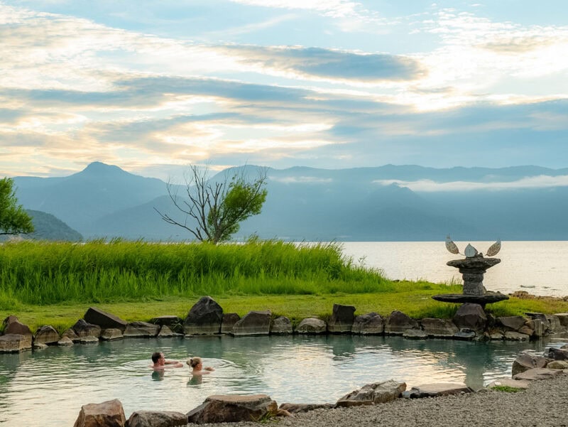 People soaking in a hot spring pool beside a lake, with grassy banks and mountains silhouetted against a cloudy evening sky.