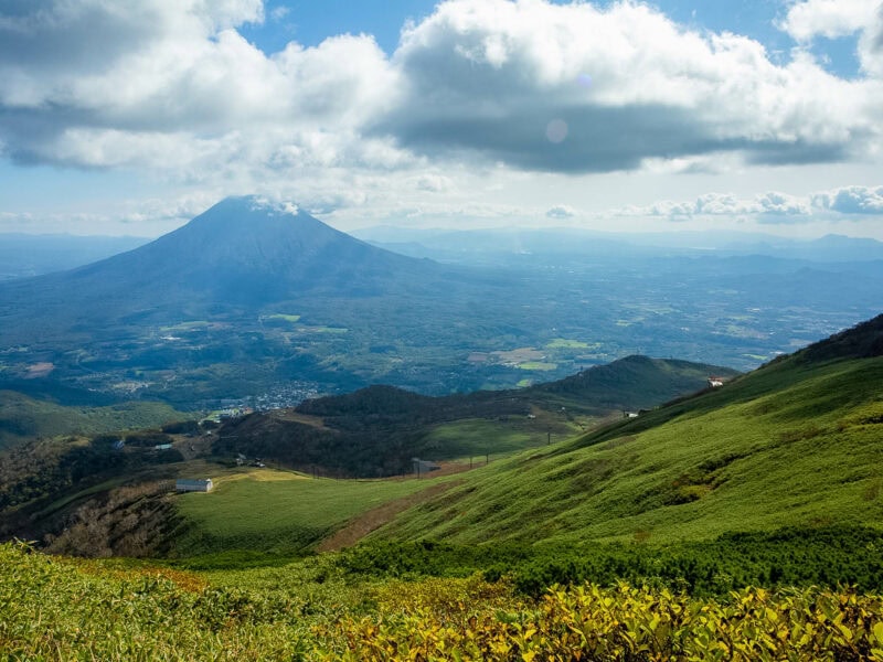 Expansive panorama of Mount Yotei with rolling green hills and patchwork farmland, beneath a partly cloudy sky.