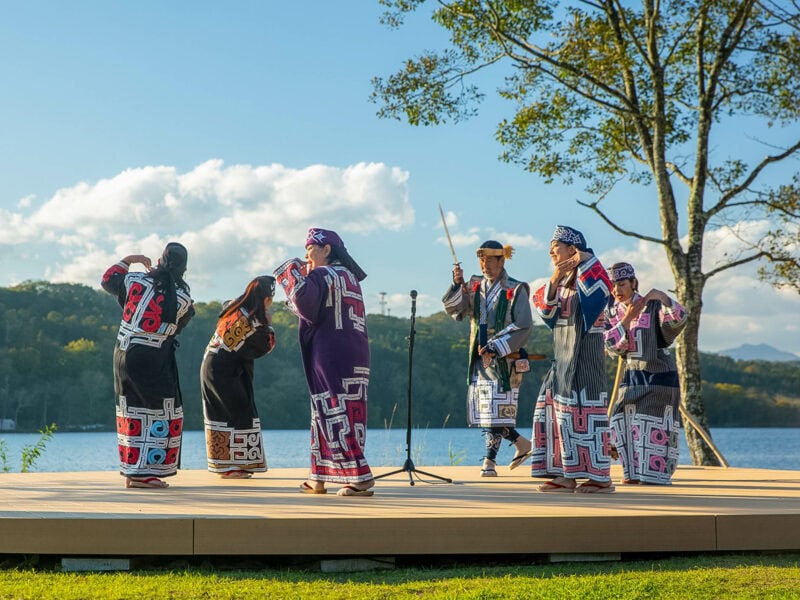 Group in patterned traditional Ainu clothing performing a dance on a wooden outdoor stage under a tree, with a lake and forest in the background and a partly cloudy blue sky.