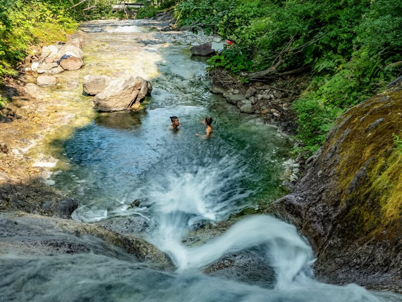 Clear river cascading over a short waterfall into a turquoise pool where two people are wading. The water then winding over boulders between steep, forested banks
