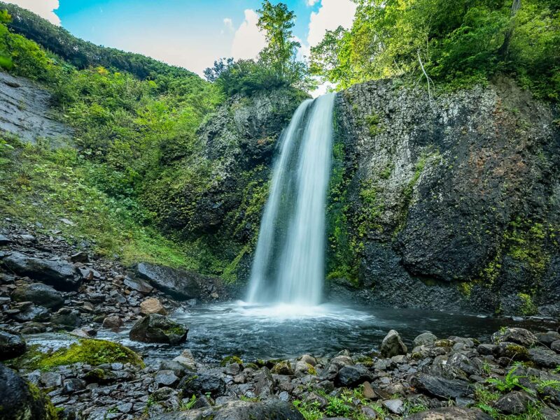Tall waterfall cascading into a small rocky pond surrounded by forested cliffs and a blue cloudy sky in the background.