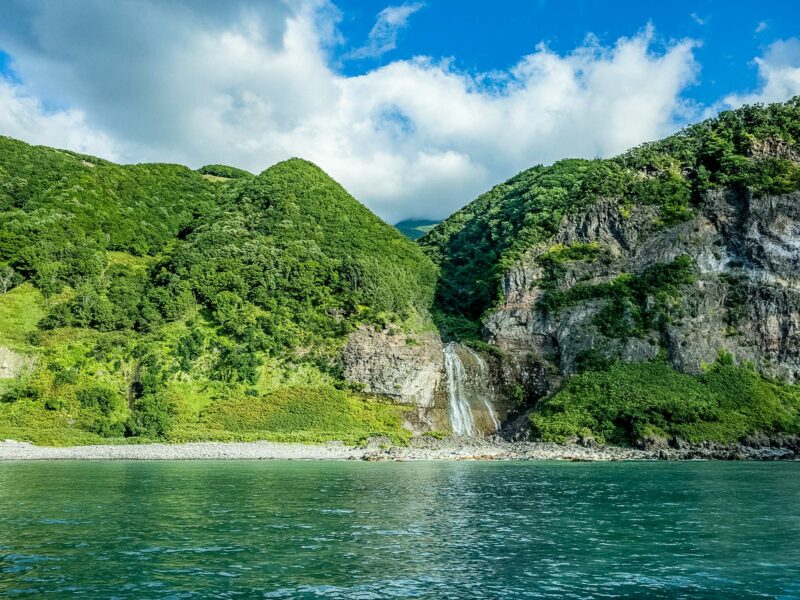 Small, multi-tier waterfall dropping from lush, green cliffs to a narrow pebble shore, viewed from calm turquoise sea under patchy white clouds.