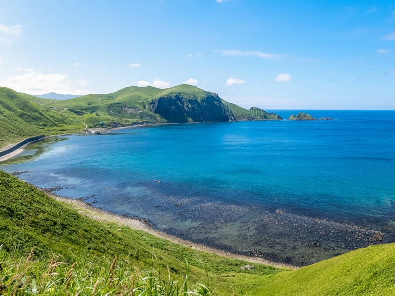 Turquoise bay with a curved grassy headland and low rocky cliffs, tapering to a point with scattered offshore islets and a clear horizon.