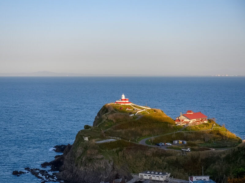 Red-roofed lighthouse perched on a rocky cliff overlooking the deep blue ocean. A shore line and some building can be seen in the hazy distance.