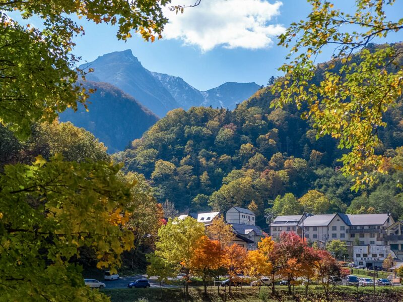 Cluster of village houses surrounded by red, orange, and yellow autumn trees, set against a backdrop of tall mountains under a partly cloudy sky.