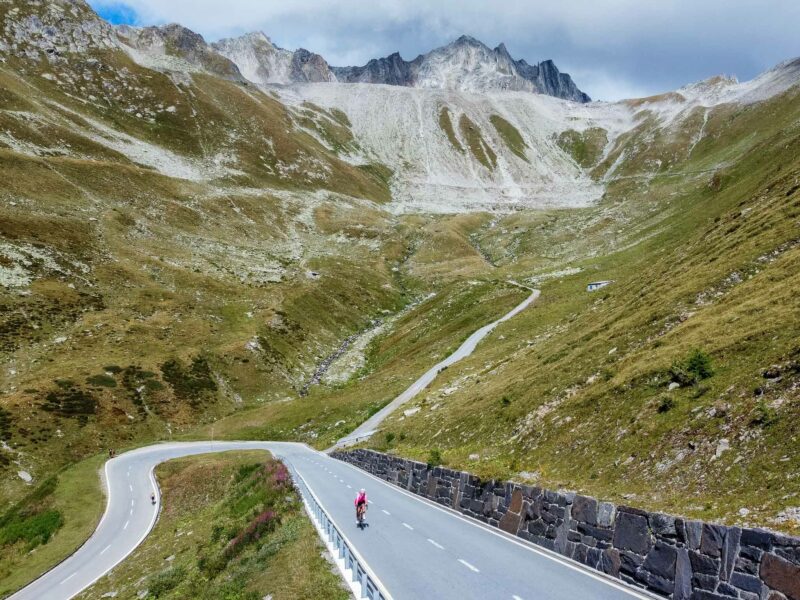 Cyclists climbing winding alpine road beneath rocky peaks