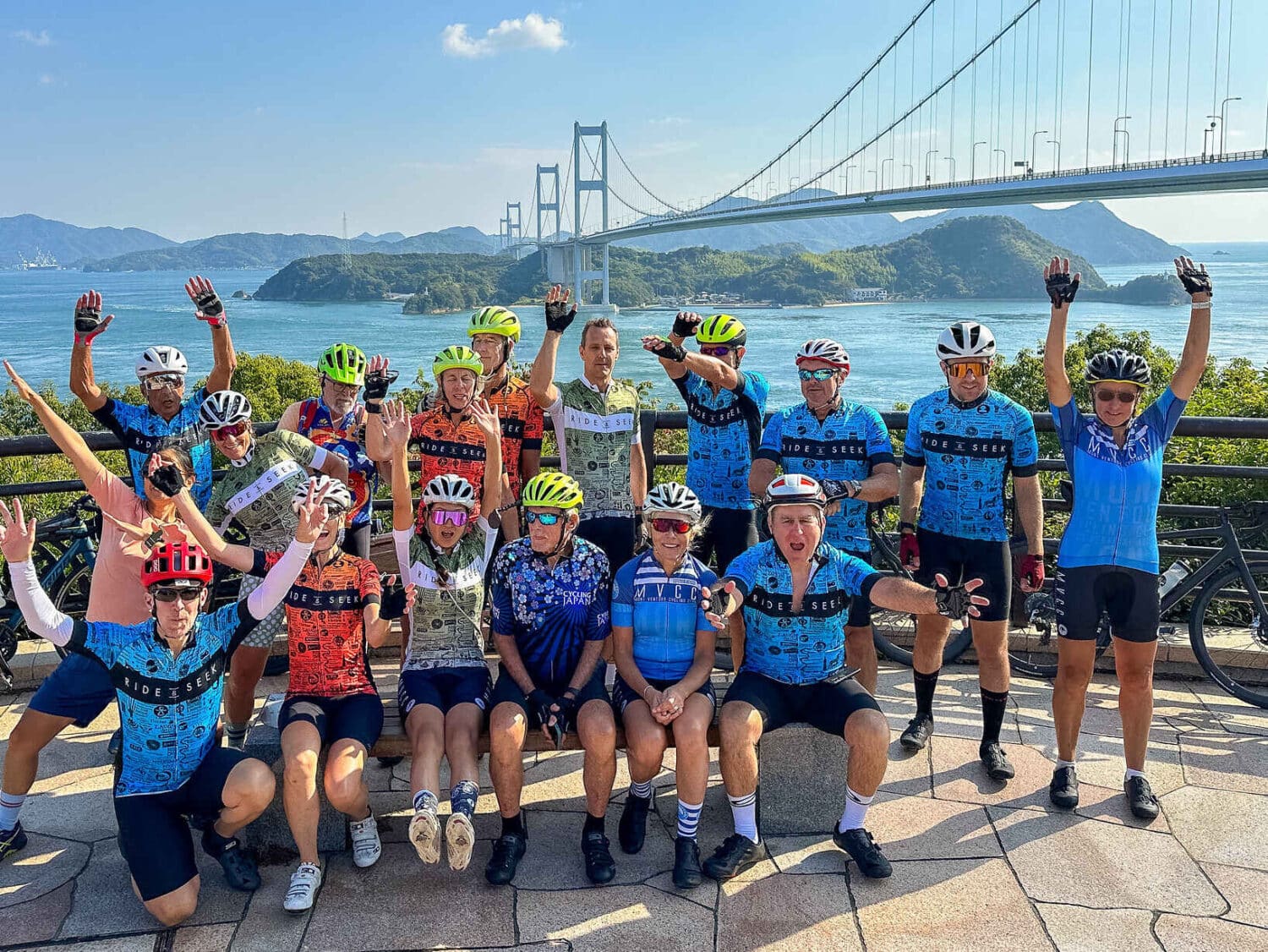 A group of cyclists posing on a scenic overlook during a Ride and Seek Japan cycling tour, featuring the Shimanami Kaido suspension bridge in the background.