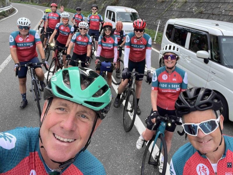 A group of cyclists wearing matching red, teal, and white "Ride & Seek" jerseys and helmets, taking a group selfie on a paved road lined with a white van and concrete retaining wall.