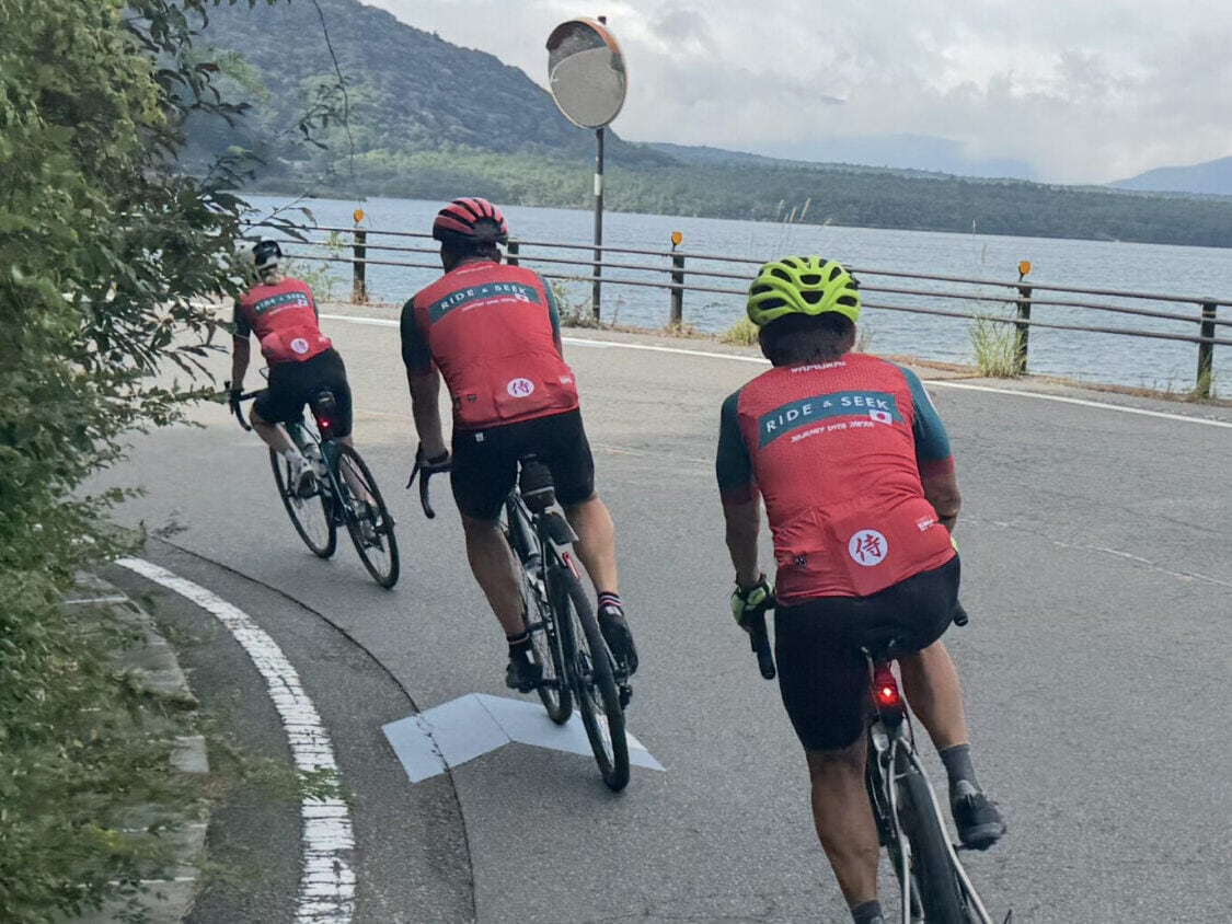 Three cyclists wearing matching red, green, and white RIDE & SEEK Samurai Collection jerseys riding along a lake. Rolling, forested hills are visible across the lake under an overcast sky.