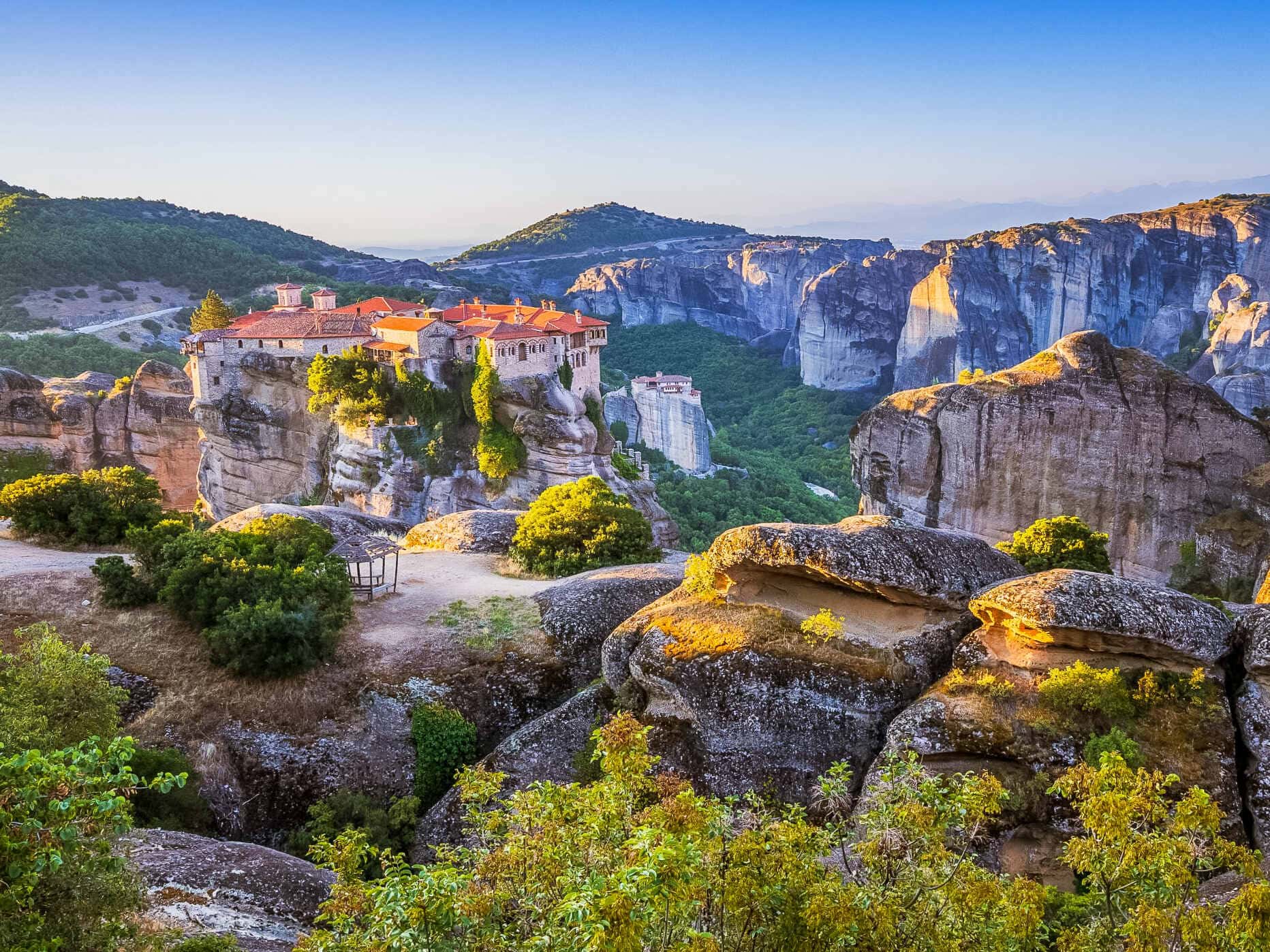 Panoramic view of Meteora in central Greece, showing a historic red-roofed Orthodox monastery perched on a sandstone rock pillar, surrounded by dramatic cliffs, forested hills and massive rock formations under a clear blue sky.